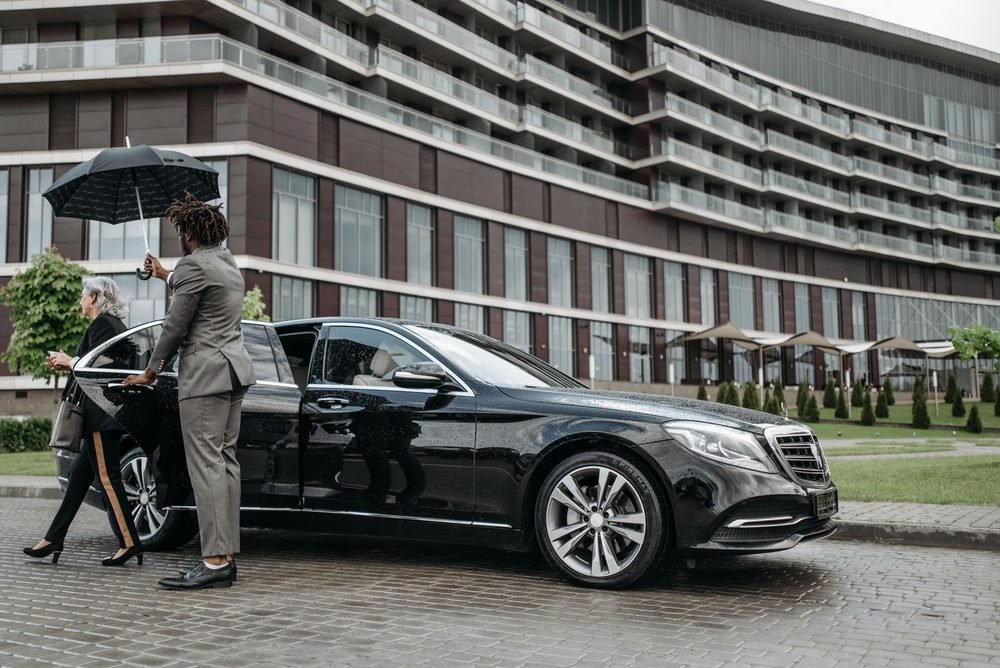 Chauffeur holding an umbrella beside a black executive sedan at a hotel portico