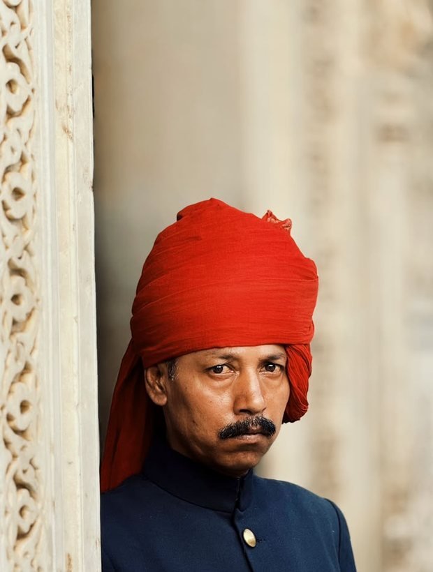 Darban in a red turban and dark tunic standing at a carved marble doorway